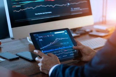 Businessman checking stock market on digital tablet and a desktop computer with stock exchange graph on screen. Financial stock market. Analyzing data in office background