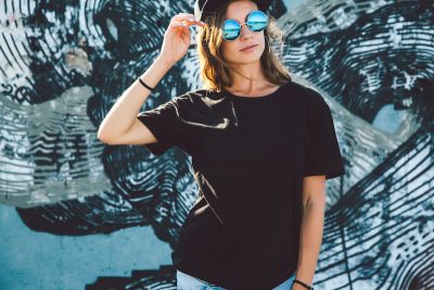 Model wearing plain black t-shirt and hipster sunglasses posing against street wall, teen urban clothing style, mockup for tshirt print store