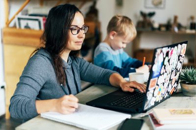 Concept of work from home. Mom and son are sitting at the desk. Business woman works on the Internet in a laptop, a child writes in a notebook.